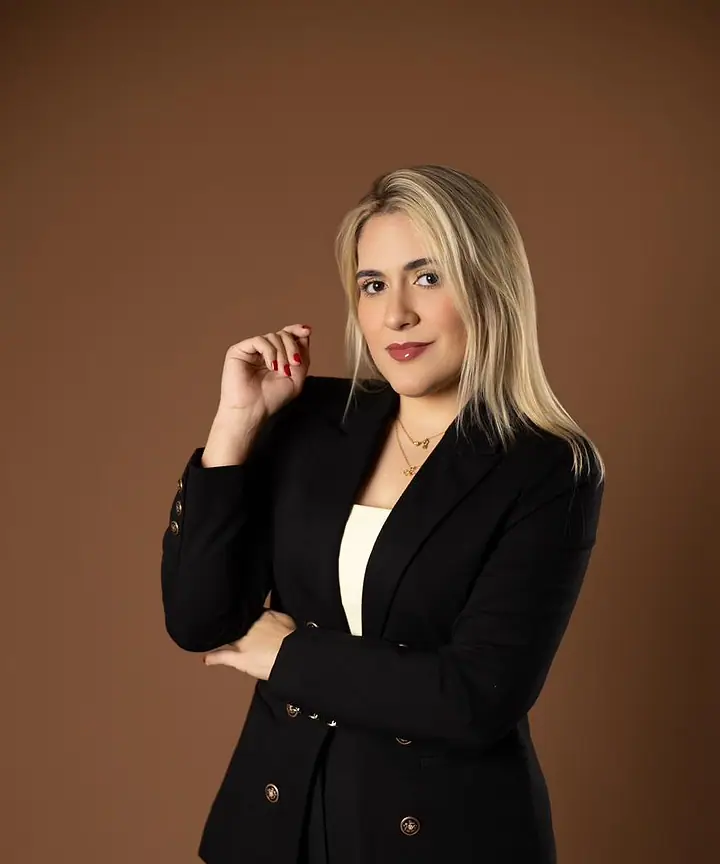 Dra. Caroline Bandeira, de blazer preto, em pé, com a mão próxima ao queixo, diante de um fundo terra